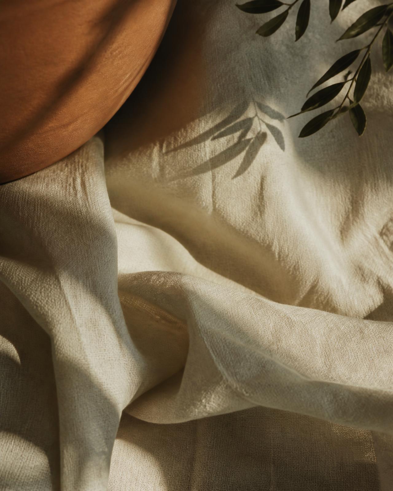 Tecido de linho claro com sombras suaves de folhas, ao lado de cerâmica em tom terracota